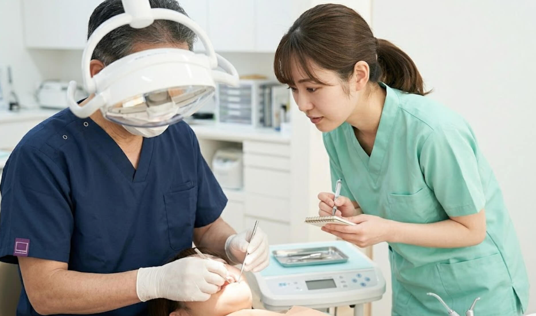 明るい歯科医院で、先輩医師の治療を熱心に見つめ、メモを取る若手女性歯科医師の後ろ姿の様子